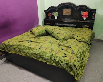 May include: A bed with a dark brown headboard and a green bedspread and matching pillows. The bedspread has a repeating pattern of black and red leaf-like shapes. The headboard has decorative carvings and small potted plants.