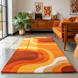 Könnte beinhalten: Ein rechteckiger Teppich im Retro-Stil mit einem wellenförmigen Muster in Orange-, Gelb- und Cremetönen. Der Teppich liegt auf einem grauen Boden in einem Wohnzimmer. Das Design erinnert an die 1970er Jahre, mit dem Text "KOHEETOORUGS" sichtbar.