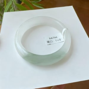 May include: A translucent jade bangle bracelet with a pale green hue. The smooth, polished surface reflects light, showcasing the natural variations in the stone. A small tag with text is attached to the bracelet. The bangle is displayed on a white surface.