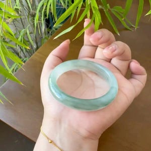 Puede incluir: Un brazalete de jade verde pálido translúcido. La superficie lisa y pulida refleja la luz, mostrando las variaciones naturales de color. El brazalete se sostiene en una mano, con una pulsera dorada visible en la muñeca. Hojas de bambú en el fondo.
