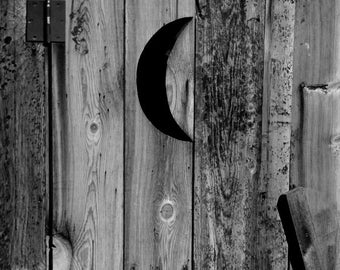 May include: A black and white photo of a wooden outhouse door with a crescent moon shape cut out. The door is made of weathered wood planks and has a metal hinge.