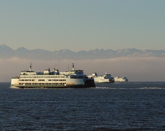 May include: Several white and green ferries sailing on a body of water under a clear blue sky. The ferries are in a line, with mountains in the background partially obscured by clouds. The water is dark blue.
