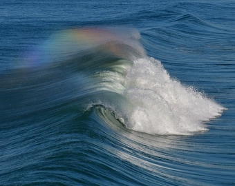 May include: A large wave breaking in the ocean with a rainbow forming in the spray.