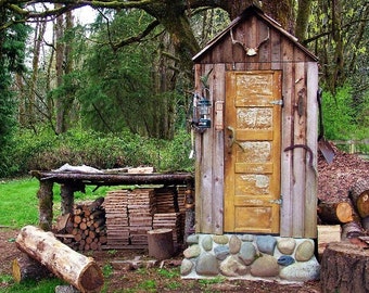 May include: A rustic wooden shed with a weathered door and a stone foundation. The shed is surrounded by trees and has a wooden shelf with stacked wood.