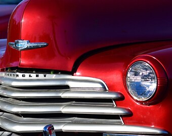 May include: Close-up of a red vintage Chevrolet car with a chrome grille and a chrome emblem. The car has a single round headlight on the right side.