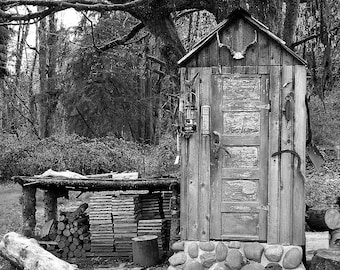 May include: A black and white photo of a small wooden outhouse with a single door. The outhouse is surrounded by trees and bushes. There is a pile of wood stacked in front of the outhouse.