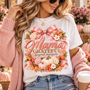 May include: A white t-shirt featuring a graphic of a pink ribbon bow, autumn leaves, and pumpkins. The text reads Mama GRATEFUL beyond measure in pink and gold lettering. The design is centered on the chest of the shirt.