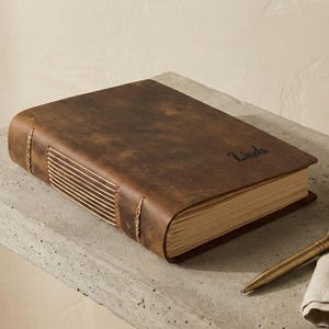 Puede incluir: Un diario de cuero marrón con el nombre "Linda" grabado en la portada. El diario está encuadernado con costuras visibles y descansa sobre una superficie de hormigón. Un bolígrafo dorado y un paño de lino están junto al diario.