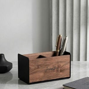 May include: A wooden desk organizer with a black metal frame. The organizer holds two pens and features the name "Jennifer" engraved on the front. A black vase and a notebook are also visible.