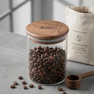 Puede incluir: Una taza de cerámica blanca con asa negra y el texto "But First, Coffee" en negrita negra. La taza está sobre un fondo borroso. La taza mide aproximadamente 10 cm de altura.