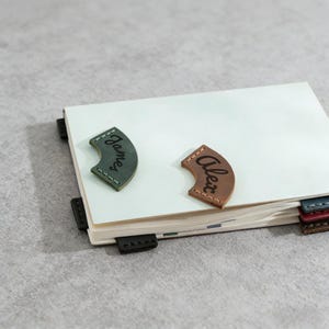 May include: A white notebook with a leather bookmark that says "James" in green and another that says "Alex" in brown. The notebook has several black, blue, and red page markers.