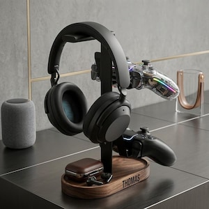 May include: A black headphone stand with a wooden base, holding black headphones, two game controllers, and a brown case. The base is engraved with the name "THOMAS". A clear controller with colorful lights is also visible.