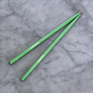 Puede incluir: Dos baquetas verdes con el nombre "Jessica" impreso en blanco. Están dispuestas en diagonal sobre una superficie de mármol. Las puntas de las baquetas son de color marrón claro.