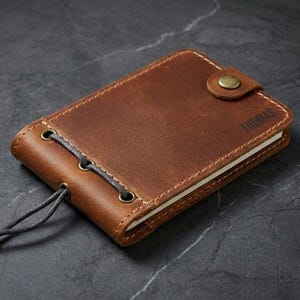 May include: A brown leather notebook with the name "THOMAS" embossed on the cover. The notebook has a button closure and a gray cord binding. The edges are stitched with a contrasting thread.