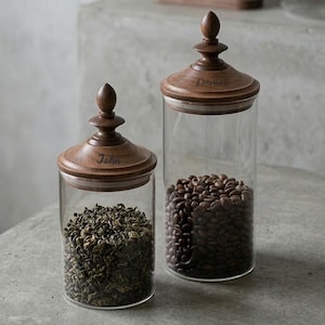Può includere: Due barattoli di vetro con coperchi in legno. Uno contiene foglie di tè verde, l'altro chicchi di caffè. I coperchi sono incisi con i nomi "John" e "Daniel". I barattoli sono cilindrici e poggiano su una superficie neutra.