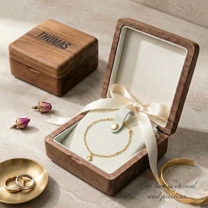 Könnte beinhalten: Eine Holzschmuckschatulle mit dem Namen "THOMAS" auf dem Deckel graviert. Im Inneren liegt ein goldenes Armband auf einem hellblauen Kissen, gebunden mit einem cremefarbenen Band. Eine kleine goldene Schale enthält zwei Ringe. Der Text "Our love is eternal and precious" ist sichtbar.