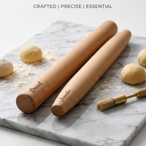 May include: Two wooden rolling pins, engraved with the names "Daniel" and "Jennifer", on a marble surface. The text "CRAFTED | PRECISE | ESSENTIAL" is at the top. A pastry brush and dough balls are also visible.