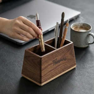 Op de afbeelding: Een houten bureau-organizer met twee compartimenten, met pennen en een penseel. Een hand plaatst een gouden en bordeauxrode vulpen in de organizer. De naam "Thomas" is gegraveerd op de voorkant. Een laptop en een mok koffie staan op de achtergrond.