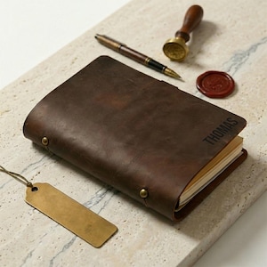 May include: A dark brown leather journal with the name "THOMAS" embossed on the cover. A brass bookmark, pen, wax seal, and stamp are arranged around the journal on a stone surface. The journal is closed and secured with brass rivets.