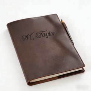 Puede incluir: Un diario de cuero marrón con el nombre "M. Taylor" grabado en la portada. Un bolígrafo negro con una funda de cuero marrón está metido en el lomo. El diario tiene un aspecto clásico y profesional.