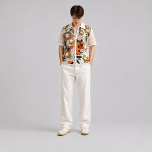 Pode incluir: Uma camisa de manga curta com bot&otilde;es, apresentando um padr&atilde;o floral de croch&ecirc; multicolorido nos pain&eacute;is frontais e uma textura de malha aberta branca nas mangas e nas costas, combinada com cal&ccedil;as brancas lisas e t&ecirc;nis brancos.