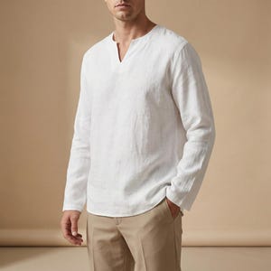 Op de afbeelding: Een wit shirt met lange mouwen en een V-hals. Het shirt is gemaakt van een lichte stof en heeft een relaxte pasvorm. Het model draagt een beige broek.