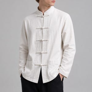 Puede incluir: Una camisa de manga larga en color blanco roto con cuello mao y cierre frontal mediante cinco botones de palanca cubiertos de tela. La prenda incluye dos bolsillos de parche en la parte inferior y está confeccionada en tejido texturizado.