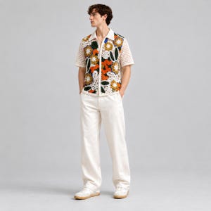 Pode incluir: Uma camisa de manga curta com bot&otilde;es em croch&eacute; cor de creme com um padr&atilde;o floral vibrante em laranja, amarelo, verde e branco. A camisa &eacute; combinada com cal&ccedil;as lisas em branco sujo e t&eacute;nis claros com solas de borracha.