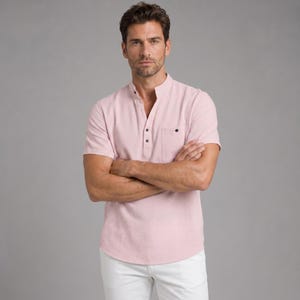 May include: A light pink short-sleeved shirt with a mandarin collar, a partial button-down placket with three visible dark buttons, and a single chest pocket on the left side. The shirt is paired with white trousers, creating a clean, neutral look.