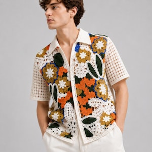 Pode incluir: Uma camisa de manga curta com bot&otilde;es e constru&ccedil;&atilde;o em croch&ecirc; com um padr&atilde;o floral multicolorido. O design incorpora fios brancos, laranja, verde-escuro e azul, com uma textura de malha aberta em forma de grade nas mangas e na gola.