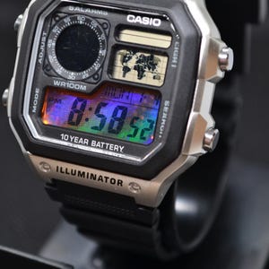 May include: A Casio digital watch with a black resin strap and a silver-tone rectangular case. The face features a world map display, a circular analog-style sub-dial, and a digital screen showing 8:58:52 with text including WORLD TIME and 10 YEAR BATTERY.