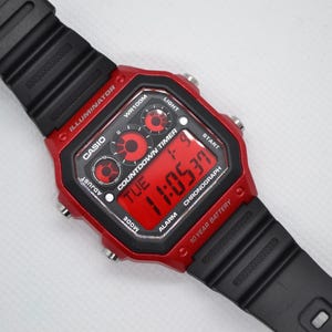 May include: A red and black Casio digital watch with a black strap. The watch face shows the time, day, and date in red digits. Features include a countdown timer, alarm, and chronograph.