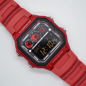 May include: A red Casio digital watch with a black face displaying the time 12:10:26. The watch has a red strap and a black and red face with the text "Casio", "Illuminator", and "Chronograph".