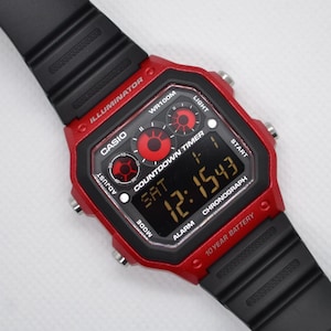 May include: A red and black Casio digital watch with a black band. The watch face displays the time, day, and various functions. The watch has the text "ILLUMINATOR", "WR100M", "COUNTDOWN TIMER", "CHRONOGRAPH", and "10 YEAR BATTERY".