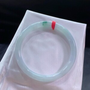May include: A pale green jadeite bangle bracelet with a hint of darker green. The smooth, polished bracelet is tied with a red string and rests on a white satin surface. The overall aesthetic is elegant and refined, with a focus on the natural stone.
