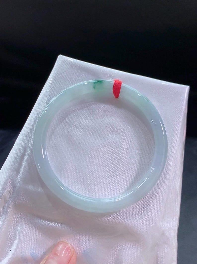 May include: A pale green jadeite bangle bracelet with a hint of darker green. The smooth, polished bracelet is tied with a red string and rests on a white satin surface. The overall aesthetic is elegant and refined, with a focus on the natural stone.