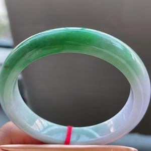 K&ouml;nnte beinhalten: Ein Jade-Armreif mit einem Farbverlauf von Gr&uuml;n nach Wei&szlig;. Der polierte Stein ist kreisf&ouml;rmig und hat eine glatte Oberfl&auml;che. Das Armband wird &uuml;ber einem gefalteten orangefarbenen Papier gehalten.