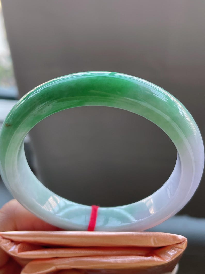 K&ouml;nnte beinhalten: Ein Jade-Armreif mit einem Farbverlauf von Gr&uuml;n nach Wei&szlig;. Der polierte Stein ist kreisf&ouml;rmig und hat eine glatte Oberfl&auml;che. Das Armband wird &uuml;ber einem gefalteten orangefarbenen Papier gehalten.