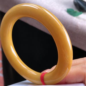 K&ouml;nnte beinhalten: Ein glattes, oval geformtes Armreif-Armband in Bernsteinfarbe. Das Armband hat eine gl&auml;nzende Oberfl&auml;che und ist einfarbig. Ein roter Faden ist um einen Teil des Armbands gewickelt. Das Armband wird von einer Person in der Hand gehalten.