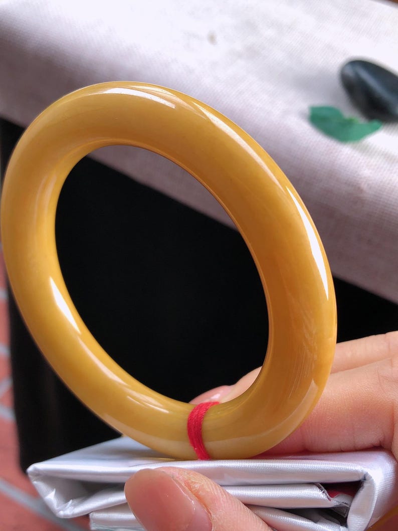 K&ouml;nnte beinhalten: Ein glattes, oval geformtes Armreif-Armband in Bernsteinfarbe. Das Armband hat eine gl&auml;nzende Oberfl&auml;che und ist einfarbig. Ein roter Faden ist um einen Teil des Armbands gewickelt. Das Armband wird von einer Person in der Hand gehalten.
