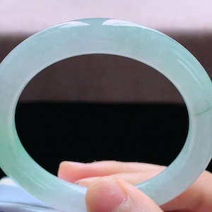 Op de afbeelding: Een lichtgroene jade armband. Het gepolijste oppervlak reflecteert licht en toont de doorschijnende kwaliteit van de steen. De armband wordt door een hand vastgehouden.