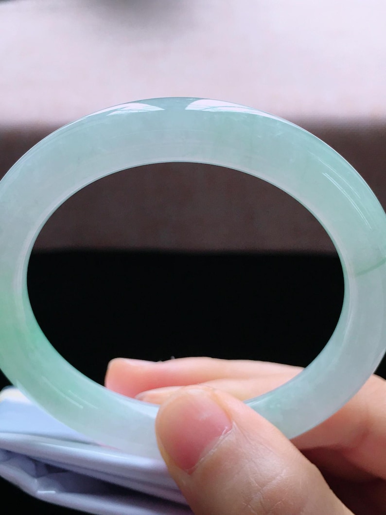 Op de afbeelding: Een lichtgroene jade armband. Het gepolijste oppervlak reflecteert licht en toont de doorschijnende kwaliteit van de steen. De armband wordt door een hand vastgehouden.