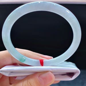 May include: A pale green jadeite bangle. The translucent bracelet has a smooth, polished finish. The jadeite is a light green colour with some darker green areas. The bracelet is held over a white satin pouch with a red string.