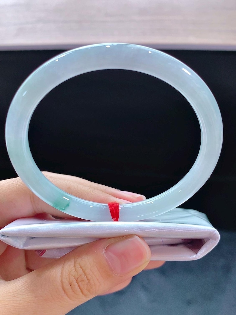 May include: A pale green jadeite bangle. The translucent bracelet has a smooth, polished finish. The jadeite is a light green colour with some darker green areas. The bracelet is held over a white satin pouch with a red string.