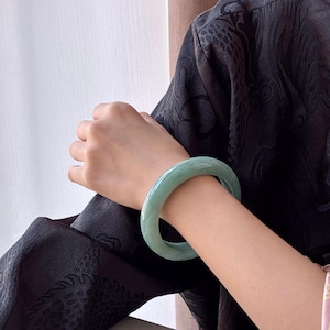 May include: A light green jade bangle bracelet. The smooth, polished bracelet is worn on a person's arm. The jade has a translucent quality, and the color is a soft, muted green. The person is wearing a black patterned top.