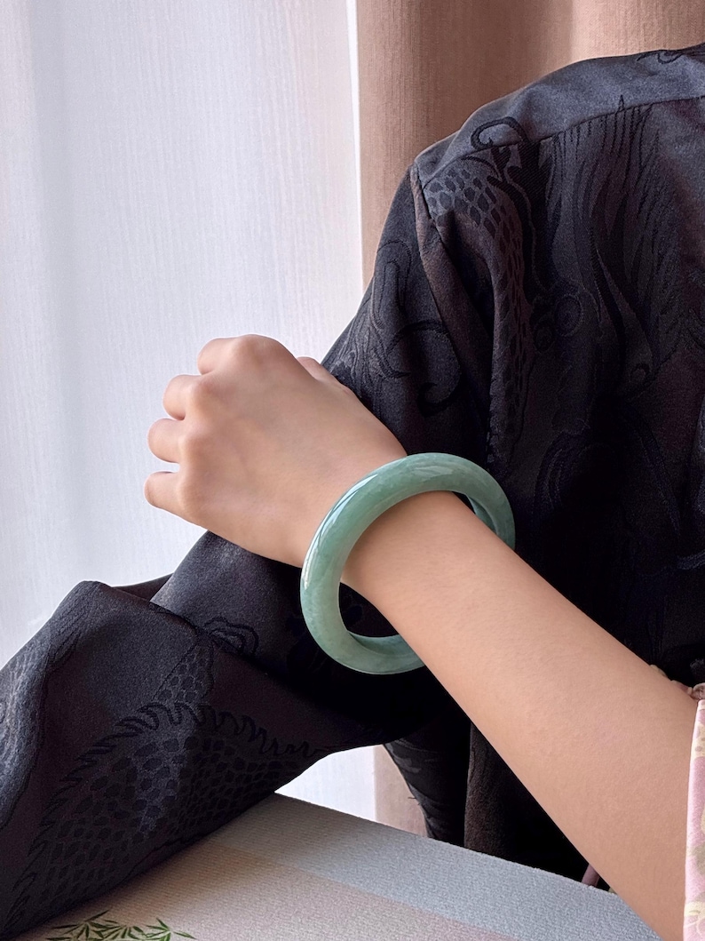 May include: A light green jade bangle bracelet. The smooth, polished bracelet is worn on a person's arm. The jade has a translucent quality, and the color is a soft, muted green. The person is wearing a black patterned top.