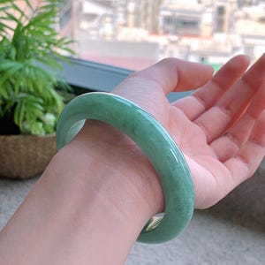 May include: A jade bangle bracelet in a light green hue. The smooth, polished surface reflects light, showcasing the natural variations in color. The bracelet is a solid, circular design, perfect for everyday wear.