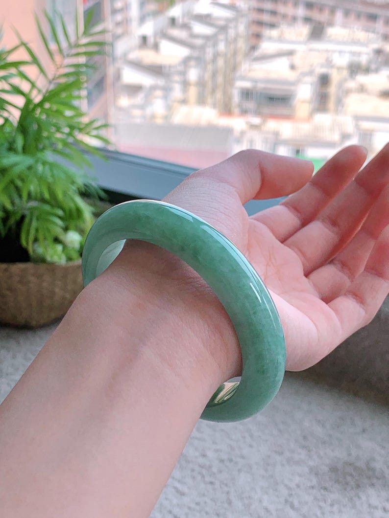 May include: A jade bangle bracelet in a light green hue. The smooth, polished surface reflects light, showcasing the natural variations in color. The bracelet is a solid, circular design, perfect for everyday wear.