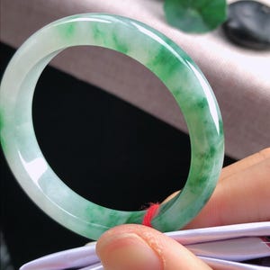 May include: A jade bangle bracelet with a translucent, light green and white marbled pattern. The polished stone is smooth and circular, with a vibrant colour gradient. The bracelet is held by a person's fingers, showcasing its natural beauty.