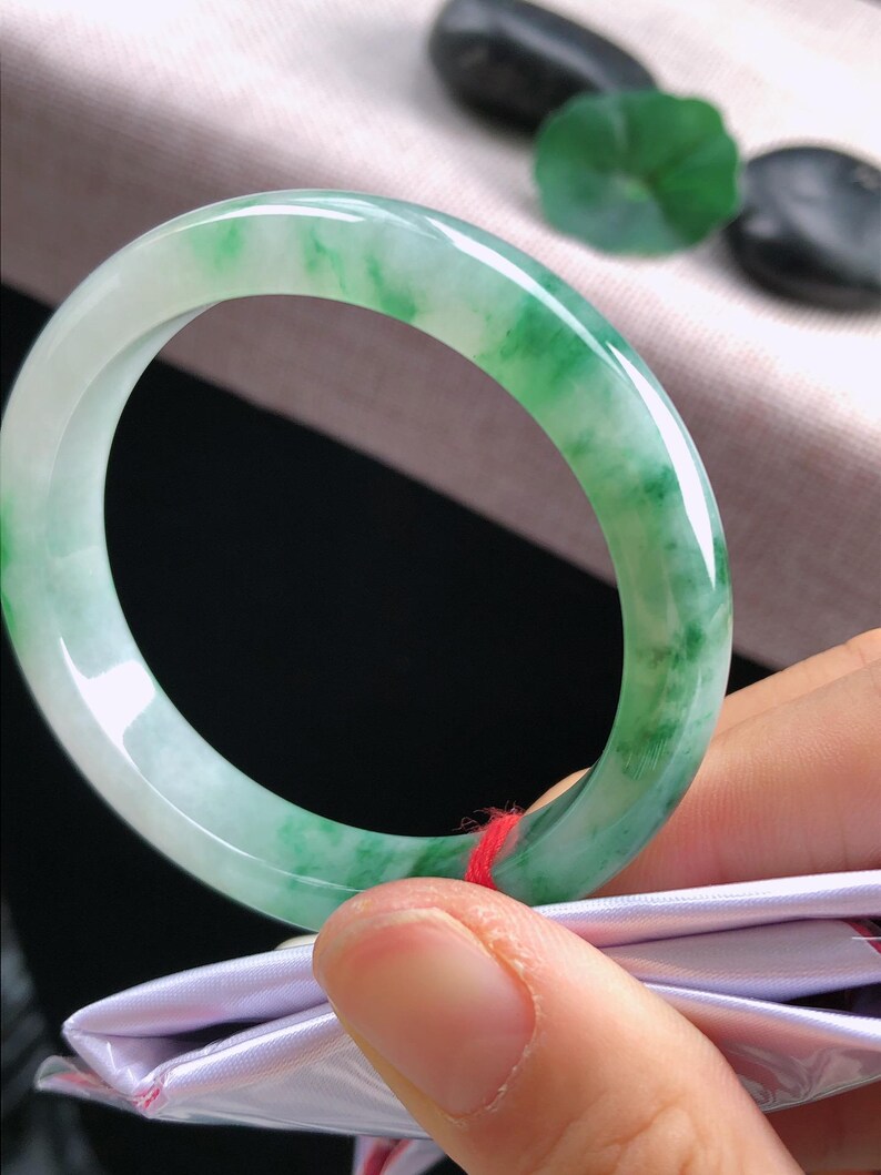 May include: A jade bangle bracelet with a translucent, light green and white marbled pattern. The polished stone is smooth and circular, with a vibrant colour gradient. The bracelet is held by a person's fingers, showcasing its natural beauty.